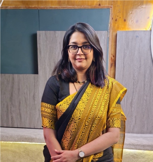 Woman in black and yellow saree with glasses standing in front of a wooden paneled wall.